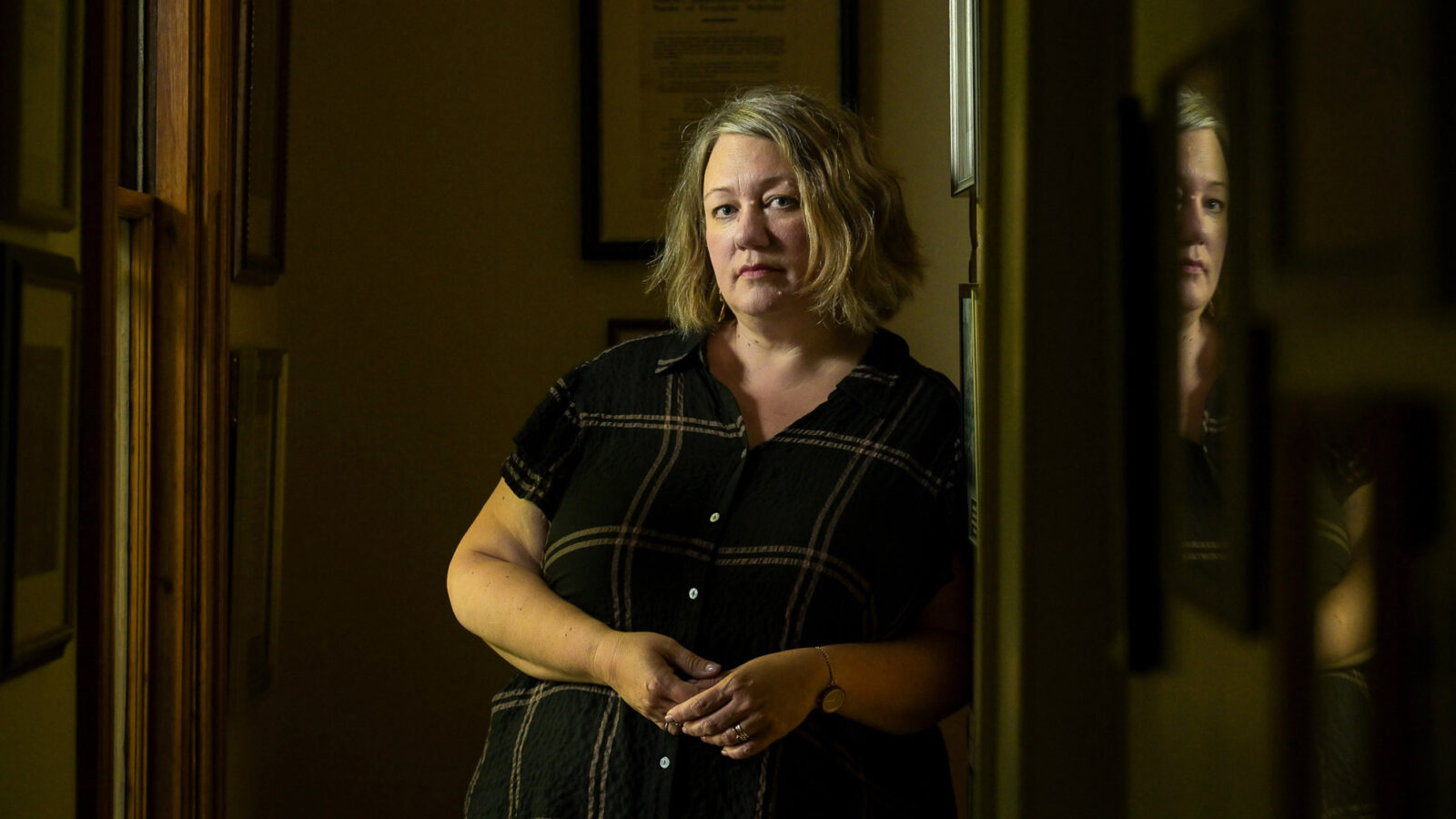 Large-bodied white woman in her mid 40s leans against a wall and stares at the camera